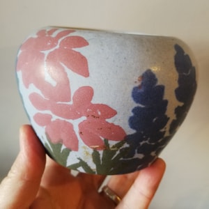 May include: A small, round ceramic vase with a light blue glaze. The vase is decorated with a floral pattern in shades of pink, blue, and green.
