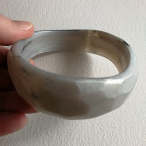Puede incluir: Una pulsera de brazalete gris claro con un diseño facetado. La pulsera tiene una superficie lisa y pulida y una forma ligeramente irregular. El color es una mezcla de gris claro y oscuro, con algunas áreas que parecen casi blancas. La pulsera está sujeta por una mano.