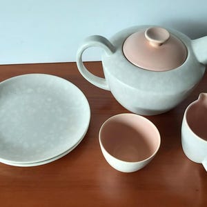 May include: A light gray ceramic tea set with a pink interior. The set includes a teapot, creamer, sugar bowl, and two plates. The teapot has a rounded shape with a matching pink lid. The creamer has a handle.