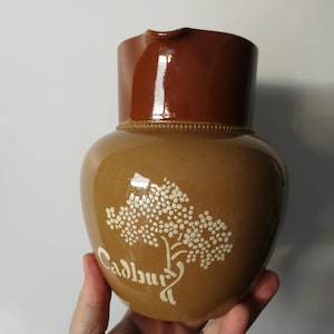 Könnte beinhalten: Brauner Keramikkrug mit dunkelbraunem Deckel und weißem Baumdesign. Das Wort "Cadbury" ist in Schreibschrift unter dem Baum geschrieben. Der Krug wird von einer Hand gehalten.