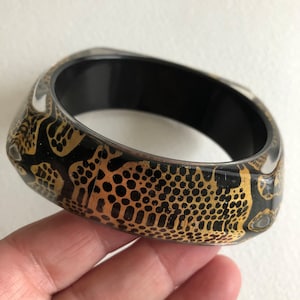 May include: Black and gold bangle bracelet with a geometric pattern. The bracelet is made of resin and has a glossy finish.