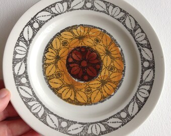 May include: A white ceramic plate with a floral design. The centre features a circular pattern of yellow and brown flowers. The rim is decorated with a grey leaf and dot pattern. A hand is holding the plate.