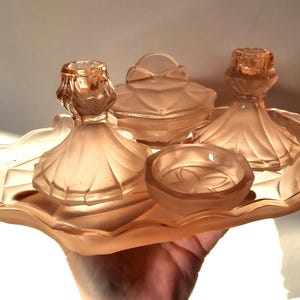 May include: A vintage, peach-colored vanity set. The set includes a tray, two candle holders, a lidded powder jar, and a small dish. The pieces have a frosted finish and a decorative, Art Deco-inspired design. The set is held in a hand.