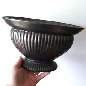 Pode incluir: Um vaso decorativo de cor bronze, com uma tigela larga e rasa e um corpo arredondado e canelado. O vaso assenta numa base circular e é segurado por uma mão. O design é clássico e elegante.