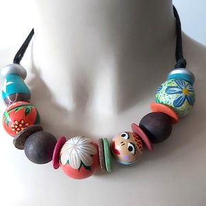 May include: A colorful beaded necklace featuring painted wooden beads in various shapes and sizes. The necklace includes beads with floral and face designs, along with brown, orange, and pink accents. The necklace is strung on a black cord.