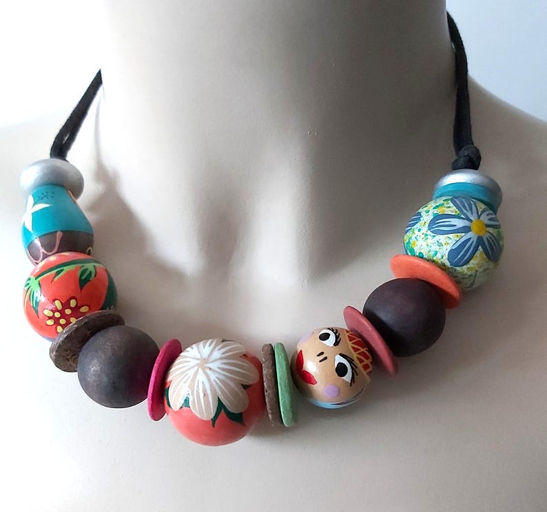 May include: A colorful beaded necklace featuring painted wooden beads in various shapes and sizes. The necklace includes beads with floral and face designs, along with brown, orange, and pink accents. The necklace is strung on a black cord.