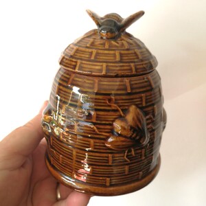 May include: A brown ceramic beehive-shaped jar with a lid, featuring a bee-shaped handle. The jar has a woven basket design with embossed bees on the sides. The jar is held by a hand.