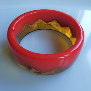Op de afbeelding: Een rode en gouden armband met een uniek ontwerp. De armband is gemaakt van plastic en heeft een glanzende afwerking.