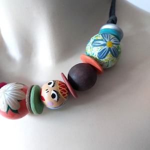 May include: A colorful, handcrafted necklace featuring painted wooden beads. The necklace includes a variety of shapes and designs, such as floral patterns, round beads, and a doll-like face. The color palette includes red, green, orange, blue, and brown.