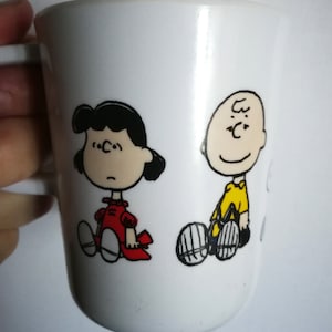 May include: White ceramic mug featuring a black and white illustration of Charlie Brown and Lucy from the Peanuts comic strip. Charlie Brown is wearing a yellow shirt and Lucy is wearing a red dress.