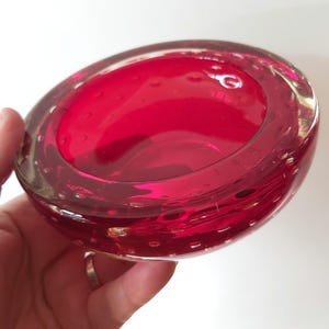 May include: A circular, deep red glass bowl featuring a thick, clear glass rim and a distinct bubble pattern embedded throughout the body. The bowl has a smooth, polished surface and a wide, open centre, showcasing the vibrant crimson colour.