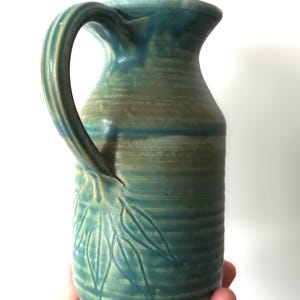 Op de afbeelding: Een keramische kan met een geglazuurde afwerking in tinten blauwgroen, blauw en aards groen. De kan heeft een geribbelde textuur langs het lichaam en een uitgesneden bladpatroon bij de basis. Het heeft een gebogen handvat en een wijde opening.