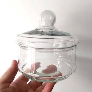 Könnte beinhalten: Ein klares Apothekerglas mit rundem Boden und passendem Deckel. Der Deckel hat einen kleinen, runden Knopf zum einfachen Anfassen. Das Glas ist zur Aufbewahrung kleiner Gegenstände bestimmt.