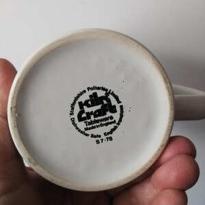 K&ouml;nnte beinhalten: Eine wei&szlig;e Keramiktasse mit Henkel, die den Boden mit einem schwarzen Stempel zeigt. Der Stempel lautet "Kiln Craft Tableware Made in England" und "Dishwasher Safe". Die Tasse wird von einer Hand gehalten.