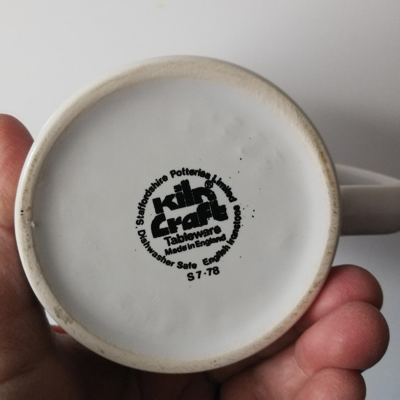K&ouml;nnte beinhalten: Eine wei&szlig;e Keramiktasse mit Henkel, die den Boden mit einem schwarzen Stempel zeigt. Der Stempel lautet "Kiln Craft Tableware Made in England" und "Dishwasher Safe". Die Tasse wird von einer Hand gehalten.