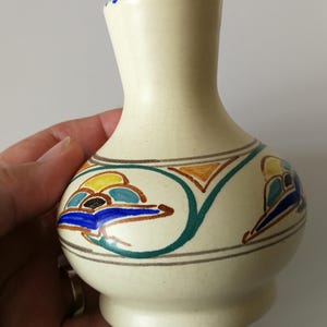 May include: Cream-coloured ceramic vase with a bulbous base and a narrow neck. The vase is decorated with a repeating pattern of colourful abstract shapes in blue, yellow, orange, and green. The rim has blue dots.