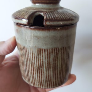 Op de afbeelding: Een cilindrische keramische pot met een bijpassend deksel voorzien van een kleine uitsparing voor een lepel. De pot heeft een tweekleurige afwerking met een glad, lichtgrijs bovendeel en een gestructureerd onderstuk met verticale bruine strepen.