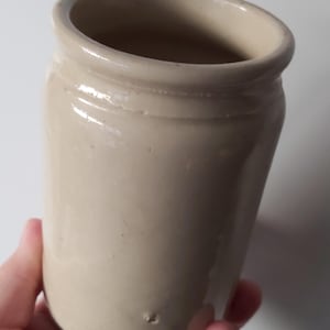 May include: A cylindrical ceramic jar with a smooth, glossy beige glaze. The vessel features a thick, rounded rim at the top and a slightly tapered body. The surface has a uniform light tan colour with a subtle texture and a small indentation near the base.