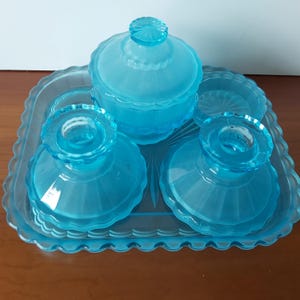 Könnte beinhalten: Ein Vintage-Waschtisch-Set aus hellblauem Glas. Das Set umfasst ein Tablett, ein Glas mit Deckel, zwei Kerzenhalter und zwei kleine Schalen. Die Teile haben einen gewellten Rand und sind auf einer Holzoberfläche angeordnet.