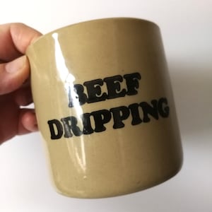 以下が含まれることがあります： 「BEEF DRIPPING」の文字が黒い太字で書かれた、ベージュ色のセラミックマグカップ。 マグカップは円筒形で、人が持っているように見えます。