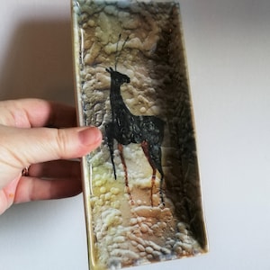 Könnte beinhalten: Eine rechteckige Keramikschale mit einer strukturierten, gesprenkelten Oberfläche in Braun-, Gelb- und Weißtönen. Auf der Schale ist eine schwarze Silhouette eines Hirsches mit langen Geweihen gemalt.
