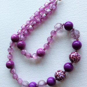 May include: A purple and pink beaded necklace with a silver clasp. The necklace features a variety of beads, including round, square, and faceted beads. The beads are arranged in a gradient pattern, with the darkest purple beads at the bottom and the lightest pink beads at the top.