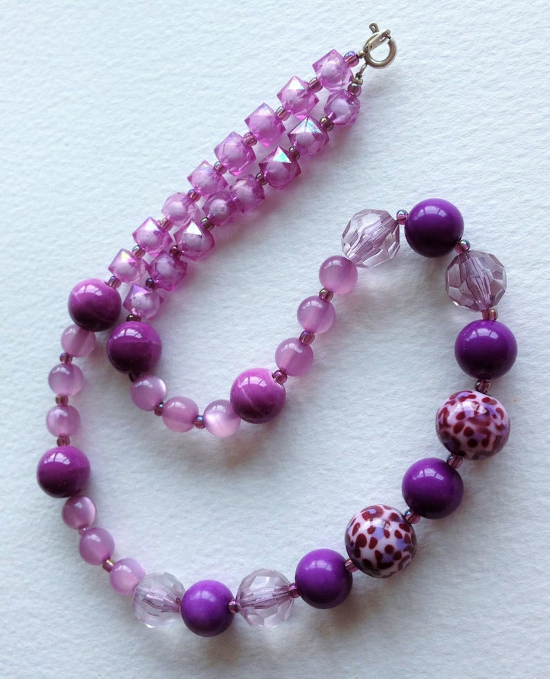 May include: A purple and pink beaded necklace with a silver clasp. The necklace features a variety of beads, including round, square, and faceted beads. The beads are arranged in a gradient pattern, with the darkest purple beads at the bottom and the lightest pink beads at the top.