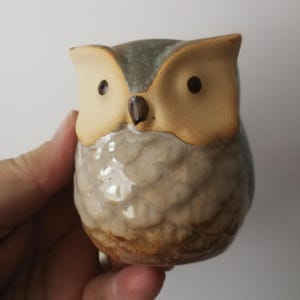 Może przedstawiać: Ceramiczna figurka sowy z teksturowanym korpusem i dwutonową kolorystyką. Sowa ma skrzydła i twarz w kolorze beżowym, z ciemnobrązowymi oczami i dziobem. Ciało to mieszanka szarości i brązu.
