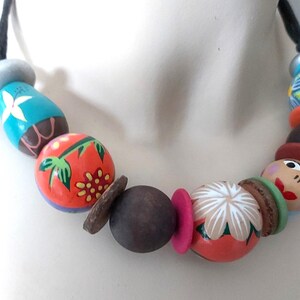 May include: A colorful beaded necklace with painted wooden beads. The necklace features a variety of shapes and sizes, including round, oval, and cylindrical beads. The beads are painted with floral and abstract designs in shades of orange, blue, and brown.