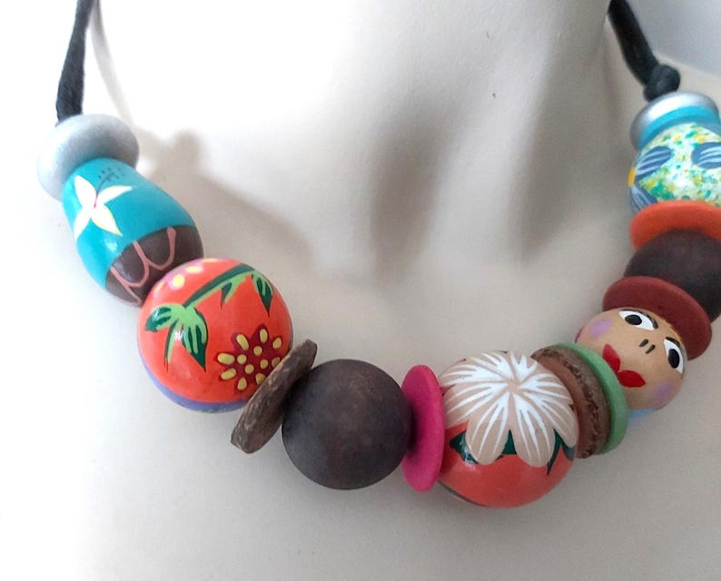 May include: A colorful beaded necklace with painted wooden beads. The necklace features a variety of shapes and sizes, including round, oval, and cylindrical beads. The beads are painted with floral and abstract designs in shades of orange, blue, and brown.