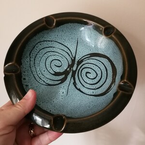 May include: A round ceramic ashtray with a dark brown rim and a light blue interior. The interior features a black spiral design. The ashtray has four small notches for resting cigarettes.