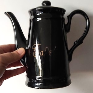 Könnte beinhalten: Eine glänzend schwarze Kaffeekanne aus Keramik mit Deckel, geschwungenem Henkel und langem Ausguss. Das Wort Café ist in goldener Schreibschrift auf der Vorderseite zu sehen. Das Design hat einen zylindrischen Körper mit einem ausgestellten Fuß.