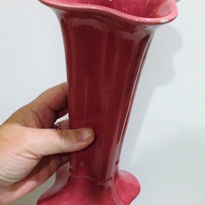 May include: A solid pink ceramic vase with a glossy finish, featuring a flared, wavy rim and a matching wide, scalloped base. The body of the vase is smooth and cylindrical, tapering slightly toward the center before widening at the bottom.
