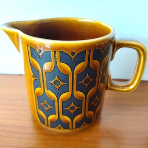 May include: A ceramic creamer jug featuring a glossy honey-brown glaze and a matte black geometric pattern. The jug has a wide pouring spout, a sturdy side handle, and a cylindrical body decorated with repeating stylised floral shapes.