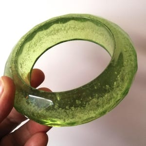 Könnte beinhalten: Ein durchscheinendes, olivgrünes Armreif-Armband. Das Armband hat eine facettierte, unregelmäßige Oberfläche und ein strukturiertes Inneres. Die hellgrüne Farbe ist durchgehend, das Stück scheint aus einem festen Material zu bestehen.