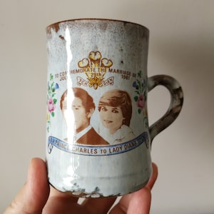 May include: A white ceramic mug with a brown glaze. It features a commemorative design for the marriage of Prince Charles and Lady Diana Spencer on 29th July 1981. The mug has a floral design and a portrait of the couple.