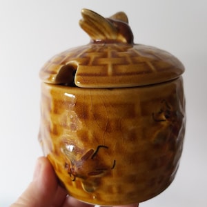 Può includere: Un vasetto per il miele in ceramica marrone chiaro con coperchio e apertura per cucchiaio. Il vasetto ha una superficie strutturata e un'ape decorativa sul coperchio. Il vasetto ha una glassa craquelé.