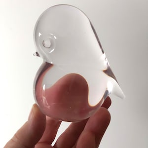 May include: A clear glass bird figurine with a rounded body and a small beak. The bird is held in a hand, and the light reflects through the glass, creating highlights and shadows. The figurine is a decorative item.