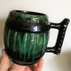 May include: A green and black ceramic mug shaped like a barrel with a black handle. The mug has a glossy finish and a ribbed design.