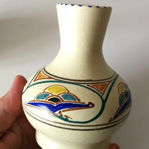 May include: Cream-colored ceramic vase with a bulbous body and a narrow neck. The vase is decorated with colorful, stylized cloud-like designs in blue, yellow, orange, and green. The rim has small blue dots.