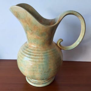 Puede incluir: Una jarra de cerámica con un esmaltado moteado en tonos verde salvia y naranja cálido. El recipiente tiene un cuerpo redondeado con detalles acanalados horizontales, un pico vertedor ancho y un asa curva que termina en un pequeño adorno.