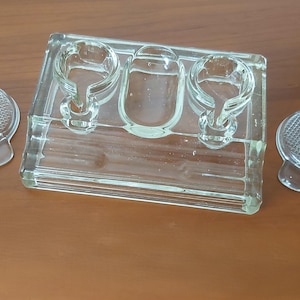 Peut inclure: Un ensemble d'encrier de bureau en verre transparent comprenant une base rectangulaire avec deux ouvertures circulaires pour les flacons d'encre, une indentation ovale centrale et deux couvercles en verre séparés avec un motif quadrillé.