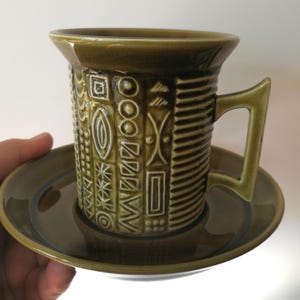Può includere: Un set di tazza e piattino da caffè in ceramica verde. La tazza presenta un motivo geometrico in bianco e poggia su un piattino abbinato. Il manico e il bordo della tazza sono di una tonalità di verde più chiara.