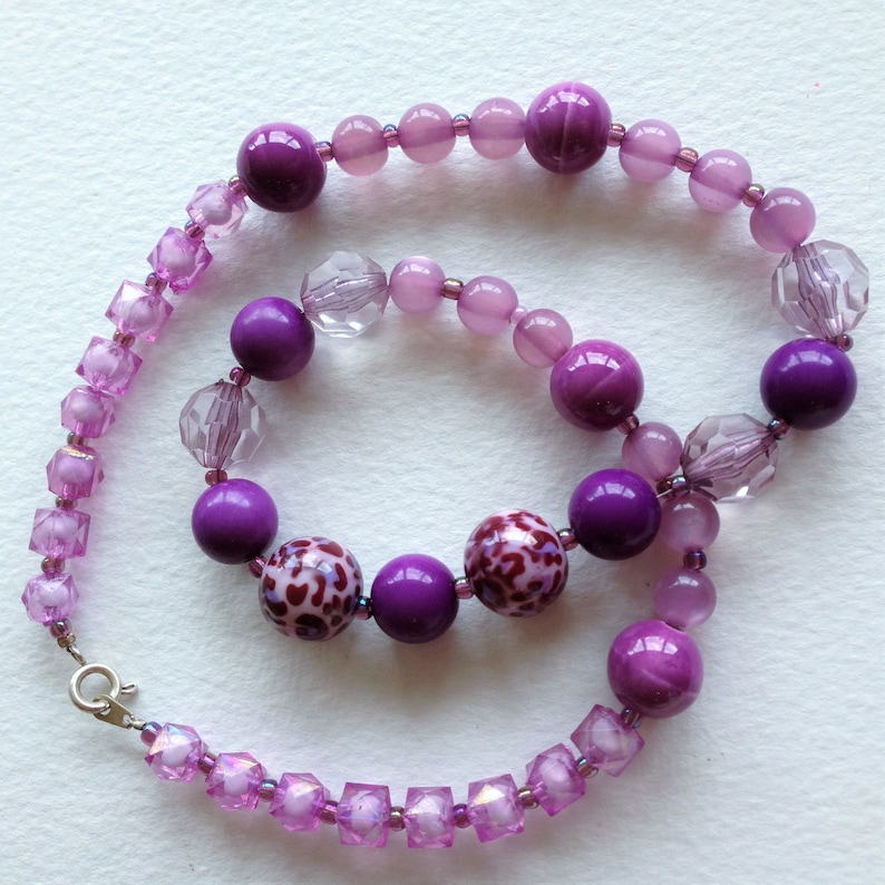 May include: A purple and clear beaded necklace with a silver clasp. The necklace features a variety of purple beads, including round, square, and faceted beads. Some of the beads have a leopard print design.