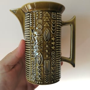 Può includere: Brocca in ceramica verde oliva con un motivo geometrico di forme bianche. La brocca ha un beccuccio, un manico e una superficie strutturata. Il design include linee, cerchi, triangoli e altre forme. La brocca è probabilmente vintage o retrò.