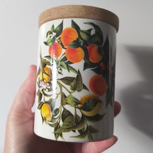 May include: A cylindrical white ceramic canister featuring a botanical print of bright orange citrus fruits, yellow lemons, dark green leaves, and small white blossoms. The container is topped with a circular, light brown textured cork lid.