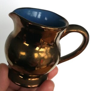 May include: A small, copper-colored ceramic creamer with a blue interior and a rounded handle.