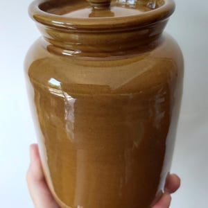 Może przedstawiać: Brązowy ceramiczny słoik z pokrywką. Słoik ma zaokrąglony korpus i lekko rozszerzony brzeg. Pokrywka ma mały uchwyt na górze. Słoik jest trzymany w dłoni.
