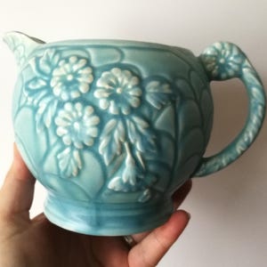 May include: A light blue ceramic creamer with a floral design. The creamer has a spout, a handle, and a round body. The floral pattern is raised and adds texture to the piece. The creamer is held in a hand.