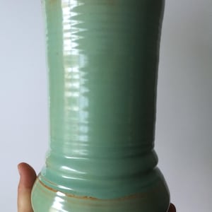 Może przedstawiać: Ręcznie robiony ceramiczny wazon w miękkim zielonym szkliwie z brązową krawędzią i podstawą. Wazon ma cylindryczny kształt z zaokrągloną podstawą i lekko rozszerzaną górą. Wazon jest trzymany przez dłoń.